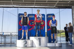Szeroki kadr: trzech młodych biegaczy z medalami na podium na scenie z widokiem na Jezioro Maltańskie.