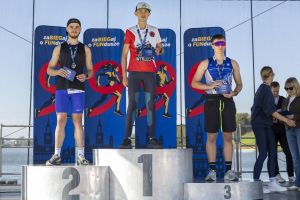 Trzech juniorów na stopniach 1–3 z medalami i niebieskimi kopertami; w tle banery i tafla jeziora.