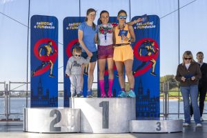 Laureatki z medalami na podium; na drugim miejscu biegaczka trzyma za rękę chłopca w szarej bluzie z nadrukiem morza.