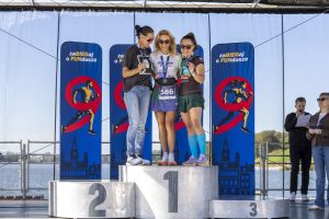 Pełne podium kobiece: trzy medalistki w okularach słonecznych, w tle woda Malti i prowadzący z mikrofonem.