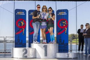 Trzy uśmiechnięte biegaczki na podium prezentują medale przed niebieskimi roll-upami zaBIEGaj o FUNdusze.