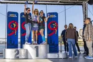 Zwyciężczynie z uniesionymi niebieskimi kopertami i medalami na podium w słoneczny dzień nad Maltą.