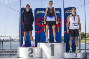 Trzech mężczyzn na stopniach podium w strojach biegowych; za nimi banery imprezy i spokojna tafla jeziora.