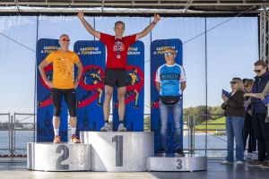 Zwycięzca w czerwonej koszulce Drużyna Szpiku na podium wznosi kciuki, obok drugie i trzecie miejsce.