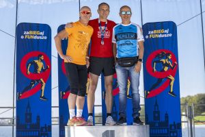 Trzech biegaczy na podium obejmuje się ramionami; środkowy z medalem, po bokach pomarańczowa i niebieska koszulka.