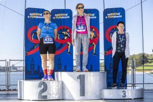 Trzy uśmiechnięte biegaczki z medalami na podium w okularach, za nimi sylwetka Poznania na banerach.