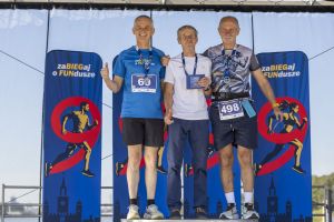 Medaliści seniorzy stoją razem na scenie z medalami; jeden pokazuje kciuk, drugi trzyma niebieską kopertę.
