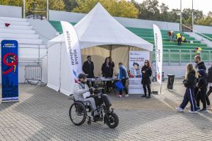 Mężczyzna na wózku z napędem elektrycznym mija biały namiot z banerem biegu i flagi Kolei Wielkopolskich.