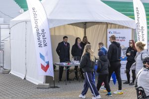 Biały namiot Koleje Wielkopolskie z obsługą w mundurach; uczestnicy biegu mijają stoisko na bruku przy trybunach.