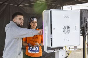 Kobieta w pomarańczowej koszulce z numerem 740 korzysta z fotokiosku pod czarnym namiotem ze studijnym światłem.