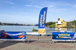 Barierki z banerami zaBIEGaj o FUNdusze nad taflą Jeziora Maltańskiego; żółta wieża finiszu regat w tle.