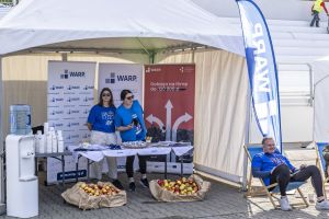 Stoisko WARP z pełnymi skrzynkami jabłek; biegacz odpoczywa w leżaku obok namiotu promocyjnego.