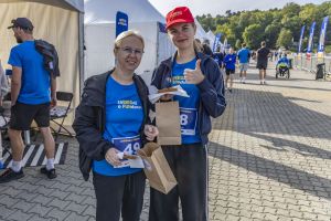 Dwie uczestniczki z przekąskami w papierowych torebkach; młodsza daje kciuk w górę w czapce z logo biegu.
