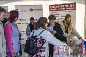 Tłum przy stoisku Wielkopolskiego Funduszu Rozwoju; plakat malowania twarzy i roll-up o historii złotówki.