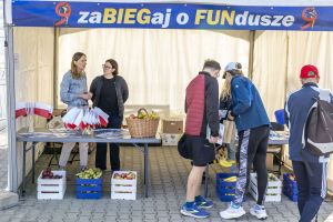Punkt z ulotkami i owocami pod banerem zaBIEGaj o FUNdusze; skrzynki z jabłkami, gruszkami i bananami.