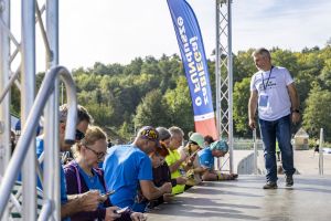 Biegacze siedzący na krawędzi sceny korzystają ze smartfonów, obok przechodzi organizator w koszulce wydarzenia.