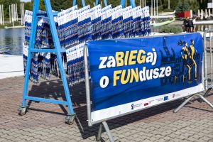 Niebieski wózek z rzędami medali finisherów stoi przy banerze zaBIEGaj o FUNdusze nad brzegiem jeziora Malta.