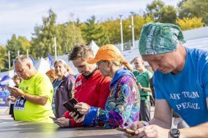 Kobieta w pomarańczowej czapce i wzorzystej kurtce sprawdza telefon w grupie zawodników przy namiotach.