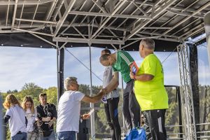 Zawodnik w zielonej koszulce Adidas wychyla się z podium, by podać dłoń organizatorowi w białej koszulce.