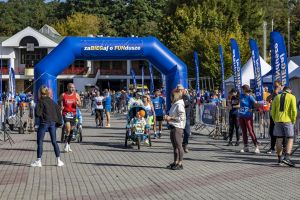 Startowiec w czerwonym topie z numerem 561 biegnie pod niebieską bramą; obok uczestnik z wózkiem biegowym.