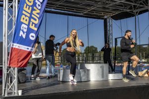 Instruktorka z mikrofonem i trener na czarnej scenie prowadzą dynamiczną rozgrzewkę przy podium i banerze.