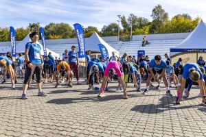 Grupa schyla się do rozciągania pleców i nóg na bruku, w centrum kobieta w różowej koszulce biegowej.