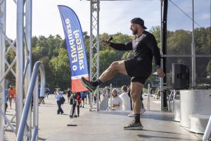 Trener w czarnym stroju z mikrofonem wykonuje wysoki kop na scenie obok niebieskiej flagi wydarzenia.