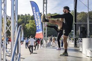 Para instruktorów na scenie demonstruje rozciąganie z wymachem nogi, w tle tłum uczestników na placu.