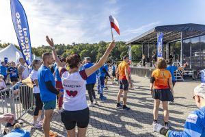 Katarzyna w białej koszulce z imieniem unosi ręce z flagą Polski przed sceną zaBIEGaj o FUNdusze na Malcie.